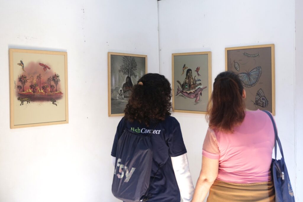 A imagem mostra duas mulheres observando obras de arte em uma galeria. As obras estão penduradas em uma parede branca e parecem ser ilustrações ou pinturas. A obra mais à esquerda é uma cena vibrante com palmeiras, animais como um felino e uma serpente, e pássaros voando em um céu alaranjado. As duas obras seguintes retratam figuras humanas em cenários mais estilizados. A primeira parece mostrar uma figura sentada sob uma árvore com outras figuras ao redor, em um estilo mais sombreado. A segunda apresenta uma figura feminina sentada em um golfinho cor-de-rosa, com pássaros e elementos que lembram tecnologia ao redor. A obra mais à direita é um estudo de borboleta, mostrando as diferentes fases de seu ciclo de vida, desde a lagarta até a borboleta adulta, incluindo a crisálida. As mulheres estão de costas para a câmera, uma vestindo uma camiseta azul escura com uma mochila nas costas, e a outra com uma camiseta rosa e uma bolsa azul. Elas parecem absortas nas obras expostas.