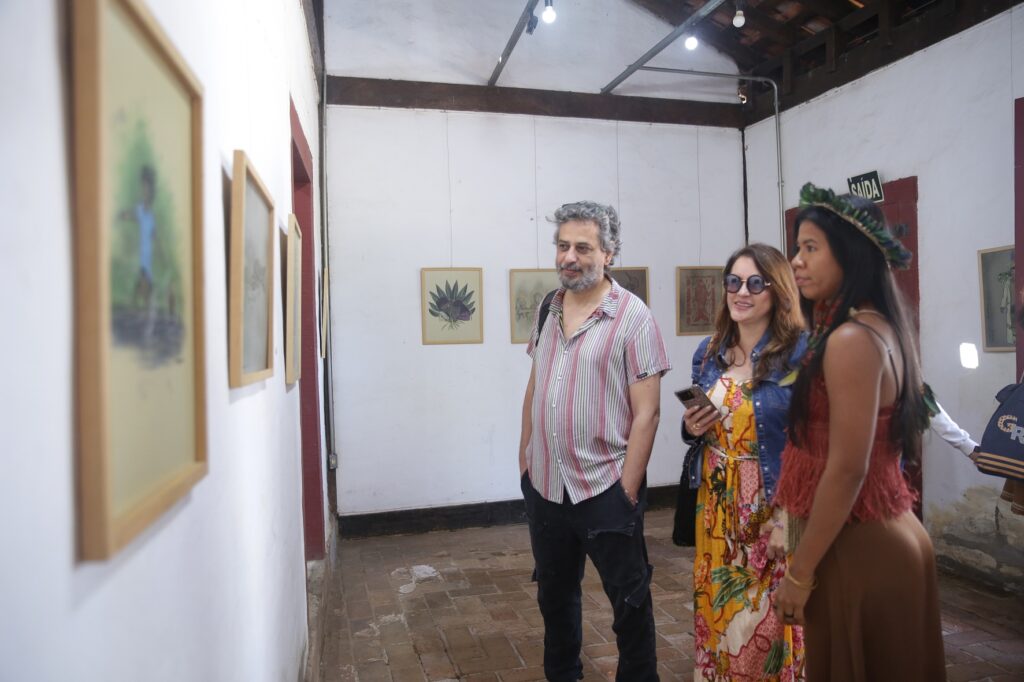 A imagem mostra três pessoas em uma galeria de arte. Um homem com cabelos grisalhos e barba está no centro, vestindo uma camisa listrada e calças pretas. Ele está olhando para uma pintura na parede. À sua direita, há uma mulher usando óculos escuros, um vestido estampado e uma jaqueta jeans. Ela está segurando um celular. Ao lado dela, outra mulher usa um chapéu decorado com penas e um vestido marrom. Ela também está olhando para a parede. As paredes da galeria são brancas e há várias pinturas penduradas, algumas delas emolduradas em madeira clara. O chão é feito de tijolos. Há uma placa de "SAÍDA" em uma porta ao fundo. A iluminação vem de lâmpadas no teto.