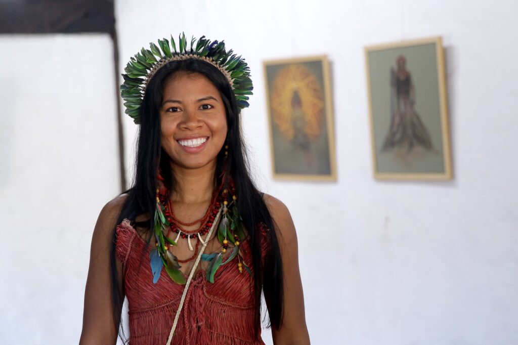 A imagem mostra uma mulher vestida com trajes tradicionais indígenas, posicionada à esquerda de um grande painel de exibição. Ela está sorrindo e segurando uma maraca levantada com a mão direita. A mulher usa um cocar feito de penas, uma blusa com franjas, uma saia longa marrom e sandálias. Uma pequena bolsa tecida está pendurada em seu ombro. O painel apresenta textos e imagens relacionadas à arte indígena. O título "Ilustração Ancestral" está em destaque em uma fonte estilizada. Abaixo, lê-se "Por Carina Horopakó". Uma seção de texto descreve Carina Desana (Horopakó Desana) como uma ilustradora do povo Desana, de Novo Airão, Amazonas, que está imersa na arte passada de geração em geração, utilizando tramas, sementes e fibras. Seu trabalho retrata a vida cotidiana de sua tribo e as lutas e conquistas dos povos indígenas do Brasil por seus direitos. O painel também apresenta uma ilustração de uma mulher indígena com um cocar de penas e um design de meia-lua. O fundo do painel é adornado com um padrão de penas e outros motivos tribais. O painel é móvel, sugerindo que pode fazer parte de uma exposição. O chão é feito de ladrilhos semelhantes a tijolos.