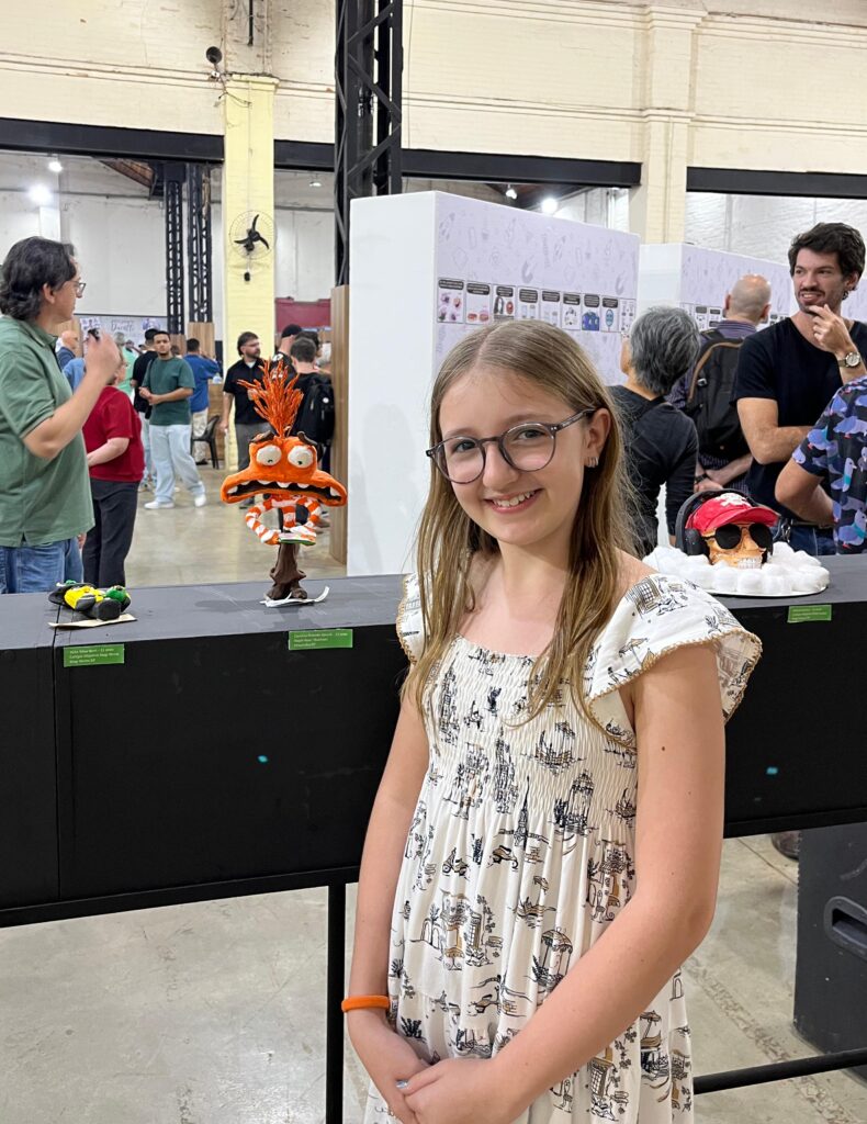 Menina de vestido claro sorri ao lado de escultura colorida em exposição no Salãozinho.