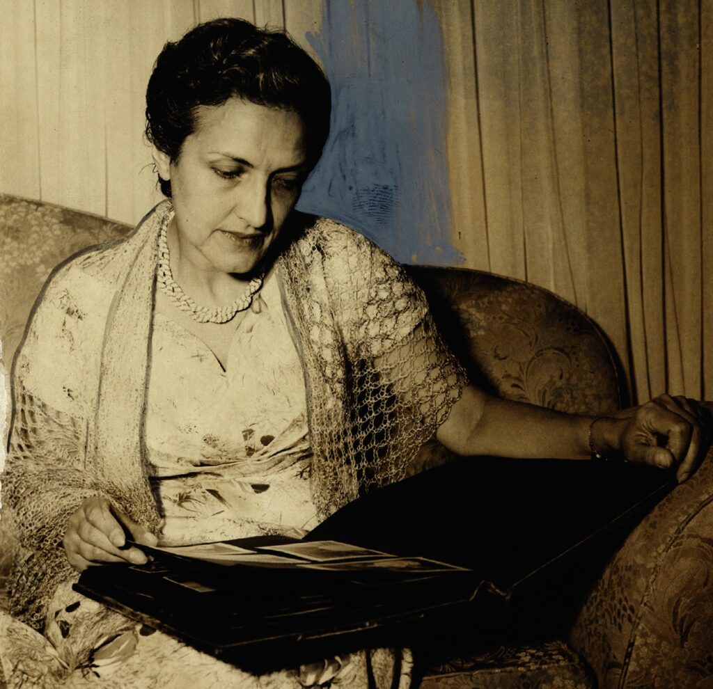 A imagem em tons de sépia mostra uma mulher sentada em uma poltrona, olhando para um álbum de fotos em seu colo. Ela usa um vestido estampado e um xale rendado sobre os ombros, além de um colar. Uma mancha de tinta azul cobre parte do seu cabelo e ombro esquerdo. As cortinas ao fundo sugerem um ambiente interno. A iluminação suave realça os detalhes do seu rosto e vestimentas, criando uma atmosfera íntima e reflexiva.