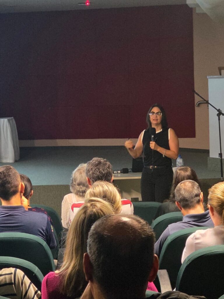 A imagem mostra uma mulher falando em um microfone em frente de pessoas sentadas em cadeiras. Ela está usando óculos, uma blusa preta sem mangas e calças pretas. A audiência está voltada para ela, e a sala parece ser um auditório ou sala de conferências. Há uma cortina vermelha ao fundo e uma mesa com um pano branco em um lado da sala. A iluminação sugere que é uma apresentação ou palestra.
