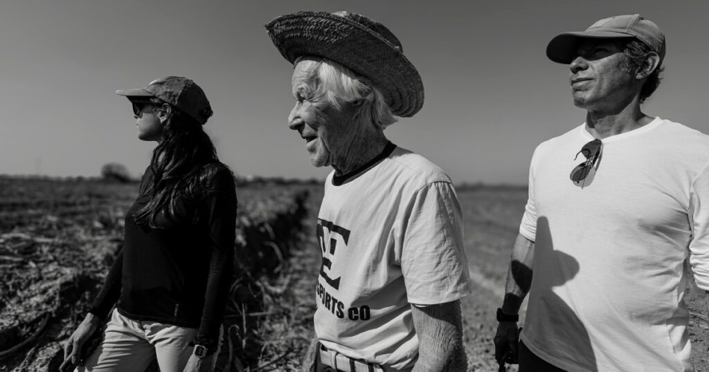 A imagem em preto e branco mostra três pessoas em um campo aberto. À esquerda, uma mulher jovem usa um boné e óculos escuros, olhando para a distância. Ela veste uma blusa preta de manga comprida e calças claras. Ao centro, um homem mais velho, com cabelos brancos e enrugado, usa um chapéu de palha e uma camiseta com uma estampa gráfica. Ele também está olhando para o lado. À direita, um homem de meia-idade, com um boné e uma camiseta branca, olha para o céu. Ele tem óculos de sol pendurados em sua camiseta. O fundo é um campo com algumas plantas mortas ou secas, e o céu está claro e sem nuvens. A luz do sol cria sombras nítidas nos rostos e corpos das pessoas.