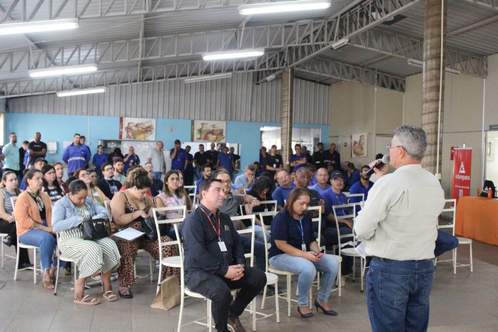 A imagem mostra um grupo de pessoas sentadas em cadeiras, assistindo a uma apresentação. Um homem mais velho, de costas para a câmera, está em pé e segurando um microfone, aparentemente falando com o público. Ele usa uma camisa clara e jeans. A plateia é composta por homens e mulheres de várias idades, a maioria vestida casualmente. Alguns estão olhando para o palestrante, outros parecem estar conversando entre si ou olhando para baixo. O ambiente parece ser um salão ou auditório, com luzes no teto e algumas decorações nas paredes, incluindo pôsteres e quadros. Há também uma bandeira vermelha com um logo no canto direito da imagem.
