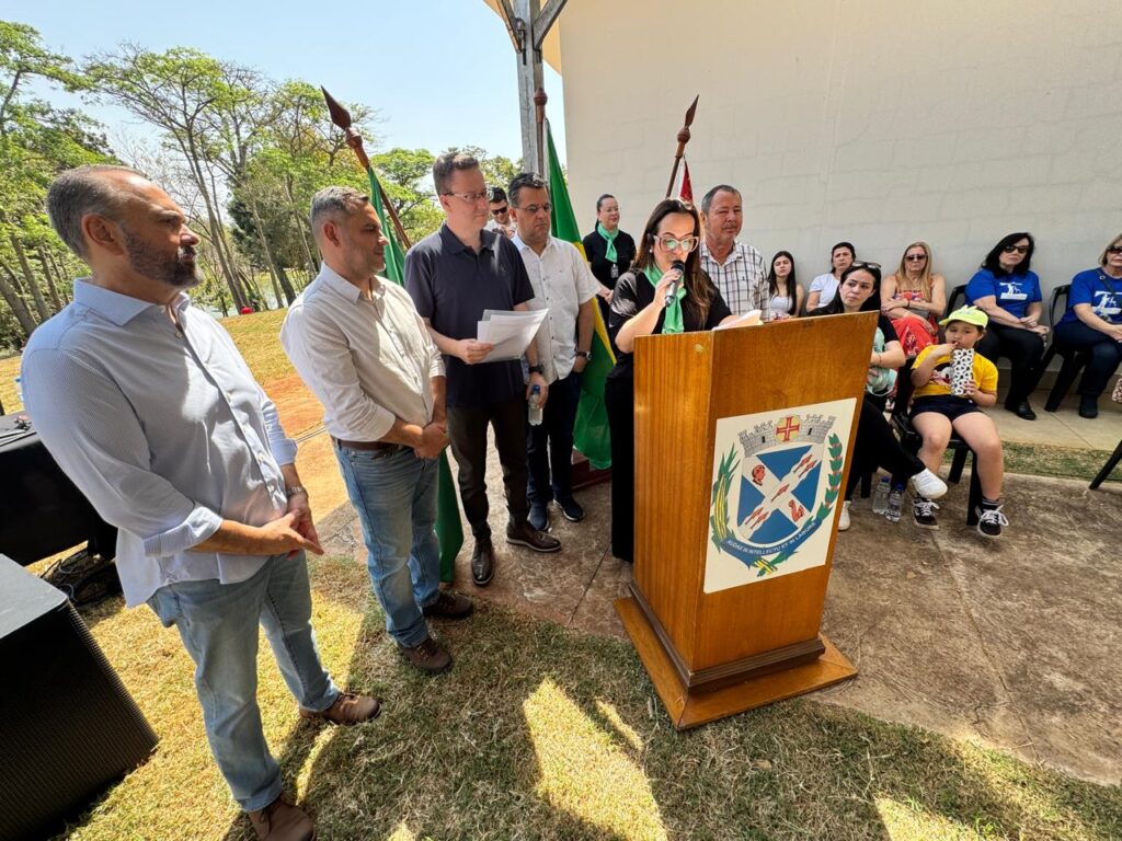 Um grupo de pessoas está reunido ao ar livre, em frente a um púlpito com um brasão. Seis pessoas estão em pé no primeiro plano; uma delas fala ao microfone, enquanto outras seguram papéis, indicando que estão fazendo discursos ou apresentações. Atrás delas, há adultos e crianças sentados, assistindo ao evento. O fundo tem uma parede branca e árvores verdes, sugerindo que estão em um parque ou área aberta.