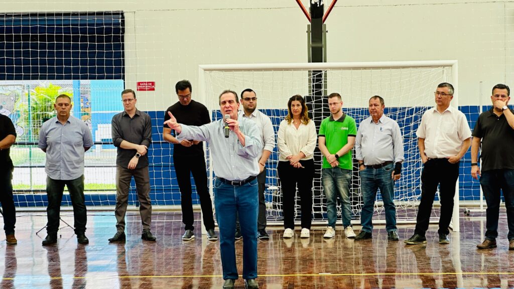 A imagem mostra um grupo de onze adultos reunidos em um ginásio esportivo, posicionados diante de uma trave de futebol com rede. O destaque vai para um homem ao centro, vestido com camisa azul clara e jeans, que fala ao microfone enquanto gesticula com a mão esquerda, indicando que está se dirigindo ao público. Os demais estão alinhados ao seu lado, com trajes que variam entre o casual e o social, sobre um piso de madeira polida.