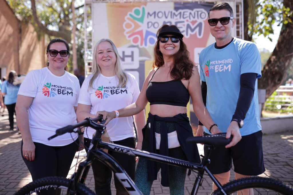 Quatro pessoas estão em pé, sorrindo, com uma bicicleta à frente delas. A mulher mais à esquerda usa uma camiseta branca com o logotipo "MOVIMENTO DO BEM" e óculos escuros. Ao lado dela, outra mulher, também com uma camiseta branca com o mesmo logotipo, usa um colar. A terceira pessoa, uma mulher, veste um top preto e leggings com estampa degradê, um boné e óculos escuros. Ela está com a mão no guidão da bicicleta. O homem à direita usa uma camiseta azul clara com o logotipo "MOVIMENTO DO BEM", óculos escuros e calção. Ele está com a mão no selim da bicicleta. Todos parecem estar em um evento ao ar livre, com uma faixa com o logotipo "MOVIMENTO DO BEM" ao fundo. O chão é pavimentado com pedras e há árvores ao redor.