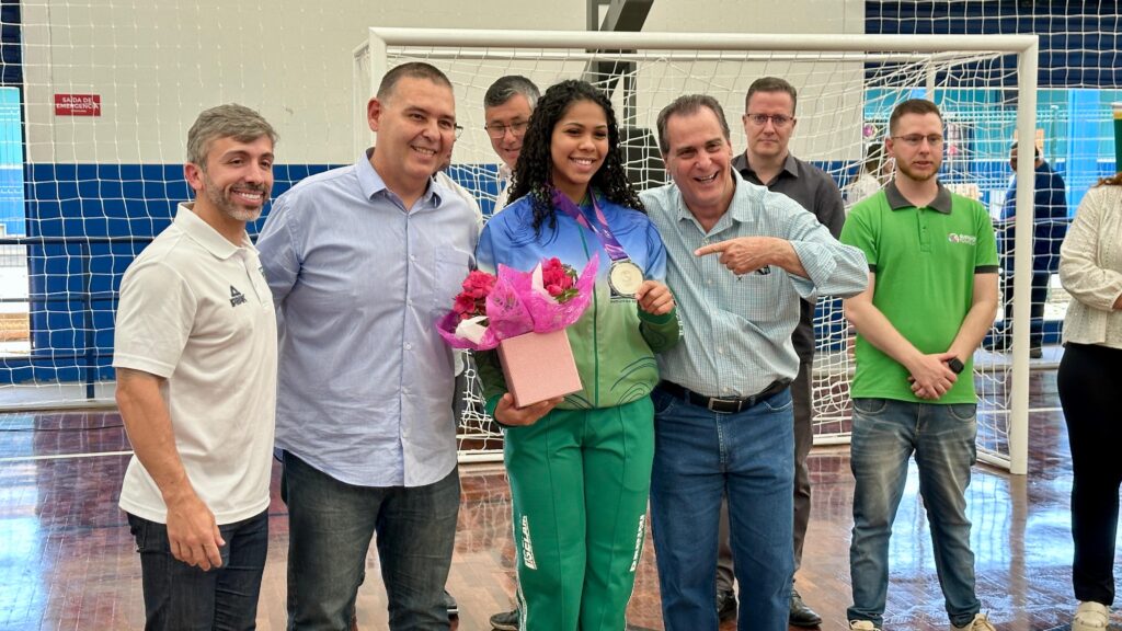 A mulher no centro, segurando um pequeno arranjo de flores vermelhas, está vestida com um vestido listrado em preto e branco. Ela está cercada por vários homens em diferentes trajes casuais e semi-formais. A iluminação sugere que a foto foi tirada em um ambiente bem iluminado, possivelmente com luzes de teto. A maioria das pessoas está sorrindo e olhando para a câmera, indicando um momento de celebração ou reunião.