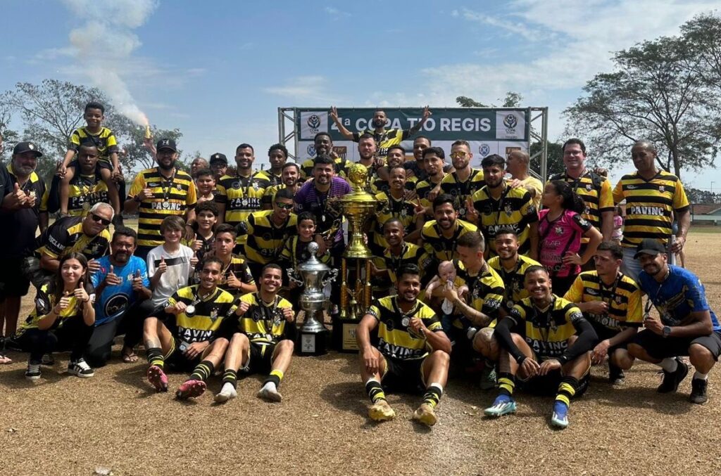 A imagem exibe um time de futebol posando para uma foto após uma partida, comemorando uma vitória. O grupo, composto por homens e algumas crianças, veste uniformes listrados em amarelo e preto, com a inscrição "PAVING" em alguns deles. Eles estão em campo, com uma faixa ao fundo que anuncia "A COPA TIO REGIS". Vários membros do time seguram troféus, medalhas e soltam fogos de artifício, evidenciando a celebração. Um bebê no colo de um dos jogadores adiciona um toque familiar à cena. O clima é de alegria e conquista, com muitos sorrisos e gestos de comemoração.