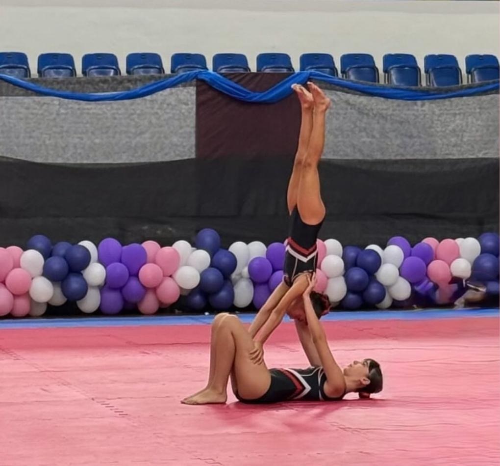 Na imagem aparece duas ginastas realizando acrobacias. A mais velha segura a mais nova que está de cabeça para baixo, num movimento sincronizado. O tablado do ginásio é de cor rosa. Ao lado das ginastas aparecem alguns balões roxo, branco e rosa.