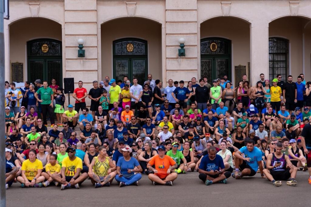 A imagem mostra um grande grupo de pessoas sentadas em degraus em frente a um edifício com arquitetura clássica. A maioria das pessoas está vestida com roupas esportivas, sugerindo que podem estar participando de um evento de corrida ou caminhada. O grupo é diversificado em termos de idade e gênero, com homens e mulheres de várias etnias. As pessoas estão sentadas em diferentes níveis dos degraus, com algumas em pé ao fundo. A iluminação sugere que é dia, possivelmente meio-dia ou início da tarde, com o sol iluminando a cena de forma uniforme. O edifício ao fundo tem uma fachada de pedra clara com arcos e janelas escuras. Há uma música tocando em um alto-falante posicionado à esquerda, e algumas pessoas parecem estar conversando entre si. A atmosfera geral é de comunidade e expectativa, como se estivessem aguardando o início de uma atividade ou o fim de um evento.