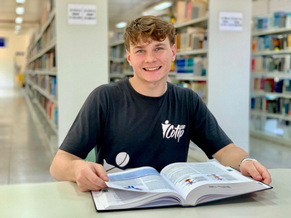 O jovem sorri diretamente para a câmera, sentado em uma biblioteca com estantes de livros ao fundo. Ele usa uma camiseta preta com um logotipo branco no peito. À sua frente, um livro grande e aberto está sobre uma mesa, e ele está folheando uma das páginas com a mão direita. A iluminação suave destaca seu rosto e o ambiente de estudo.