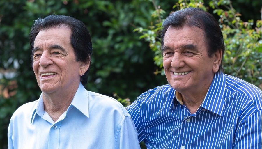 A imagem mostra dois homens mais velhos sentados lado a lado, sorrindo. O homem à esquerda usa uma camisa social azul clara e o homem à direita usa uma camisa listrada azul e branca. Ambos têm cabelo escuro e parecem estar ao ar livre, com vegetação desfocada ao fundo.