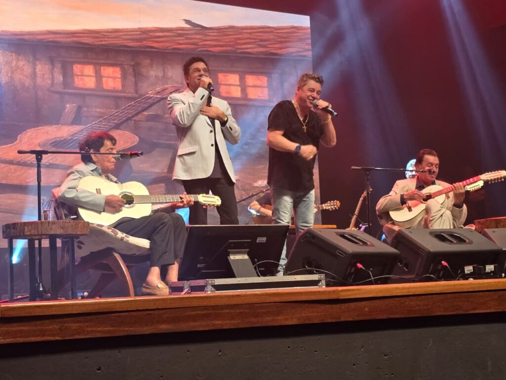 A imagem mostra quatro homens em um palco, possivelmente durante uma apresentação musical. Dois homens estão em pé no centro do palco, cantando em microfones. O homem à esquerda está vestindo um terno claro sobre uma camisa escura e tem a mão no peito. O homem à direita está vestindo uma camiseta preta com um colar e tem a mão levantada. À esquerda, um homem está sentado e tocando um violão branco. Ele está vestindo uma camisa clara e calças escuras. Há um copo de água em uma mesa de madeira ao seu lado. À direita, outro homem está sentado e tocando um violão com muitas cordas. Ele está vestindo um terno claro e uma camisa. O fundo do palco tem uma imagem de uma casa rústica com janelas iluminadas, e há mais violões emoldurados como parte da decoração. Vários holofotes iluminam o palco.