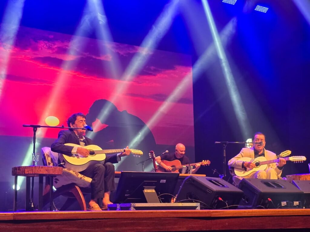 A imagem mostra três homens sentados em um palco, cada um tocando um violão. Eles estão iluminados por holofotes azuis e brancos, que criam um efeito dramático no palco. Ao fundo, uma tela exibe uma imagem de um pôr do sol com tons vibrantes de rosa e laranja, e a silhueta de uma pessoa. O homem da frente, à esquerda, usa um terno escuro e está cantando em um microfone enquanto toca seu violão. À sua direita, um homem com uma camisa clara também toca um violão. Mais atrás, um terceiro homem, careca e vestindo uma camiseta preta, toca um violão com mais intensidade. O palco parece estar em um show ou concerto, com equipamentos de som e iluminação visíveis. A atmosfera é de performance musical, com os artistas concentrados em sua música.