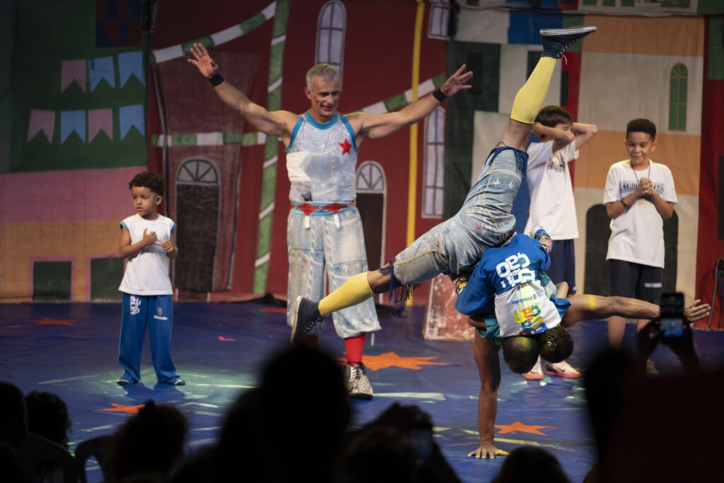 Pra cego ver: Crianças sobem ao palco e interagem com artistas circenses durante espetáculo.