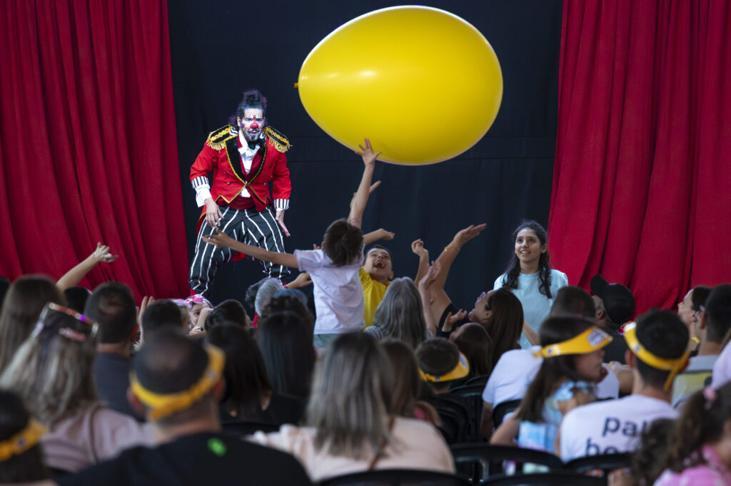 Palhaço interage com crianças jogando um balão amarelo gigante no palco.