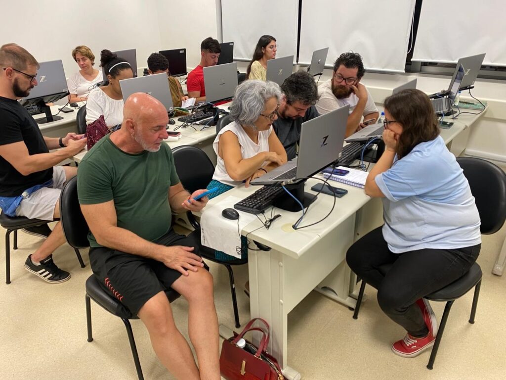 A imagem mostra um grupo de pessoas sentadas em uma sala de aula, cada uma com um laptop à sua frente. Eles parecem estar participando de uma aula ou workshop, pois todos estão focados em seus computadores. No primeiro plano, um homem careca com uma camiseta verde está sentado em uma cadeira preta, olhando para o telefone. Ao lado dele, uma mulher com óculos e uma camiseta branca está trabalhando em seu laptop. Mais atrás, outras pessoas estão sentadas em mesas semelhantes, também concentradas em seus laptops. A iluminação da sala é clara, e as persianas nas janelas sugerem que a sala está localizada em um prédio moderno. A atmosfera geral é de aprendizado e engajamento.