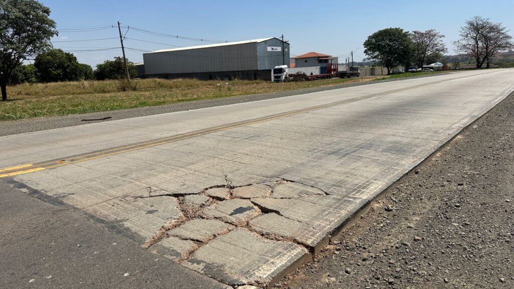 A imagem mostra uma estrada de concreto com dupla sinalização amarela no centro. No primeiro plano, há uma grande rachadura no asfalto, com pedaços de concreto soltos e quebrados. A rachadura se estende por uma área considerável da pista. Ao fundo, vemos um terreno gramado e algumas árvores. Em segundo plano, há um galpão industrial de cor cinza com um logo branco e vermelho, e ao lado, um edifício com telhado laranja. Um caminhão branco está estacionado próximo ao galpão. A estrada continua em direção ao horizonte, onde o céu azul e claro domina a paisagem.