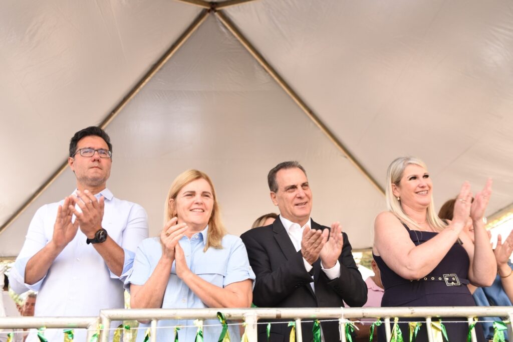 A imagem mostra quatro pessoas, duas mulheres e dois homens, em pé atrás de uma grade, aplaudindo. Eles estão sob uma tenda branca com uma estrutura de metal aparente no topo. Da esquerda para a direita, o primeiro homem usa uma camisa social branca e óculos. Ele está com as mãos juntas, como se estivesse aplaudindo. Ao seu lado, uma mulher loira, vestindo uma camisa azul clara, também aplaude com as mãos juntas. O terceiro indivíduo é um homem de terno preto e camisa branca, sorrindo e aplaudindo. A quarta pessoa é uma mulher loira com cabelo longo, usando um vestido escuro, que também está aplaudindo com as mãos juntas. Todos parecem estar em um evento ao ar livre, possivelmente uma cerimônia ou comemoração. A grade na frente deles está decorada com fitas verdes e amarelas.
