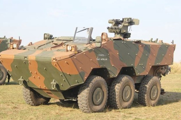 A imagem mostra um veículo blindado de transporte de pessoal (APC) 8x8, camuflado em tons de verde, marrom e bege. O veículo tem um design angular e possui seis rodas visíveis em cada lado, com a frente do veículo inclinada para cima. No topo do veículo, há uma torre equipada com uma metralhadora e possivelmente um lançador de mísseis. Há também um para-brisa com um limpador de para-brisa visível. A lateral do veículo exibe uma camuflagem em padrão de listras irregulares. Na lateral, há texto em branco que parece ser "1º BTL" e "TAH APOLLO". O veículo está estacionado em um campo gramado sob um céu claro.