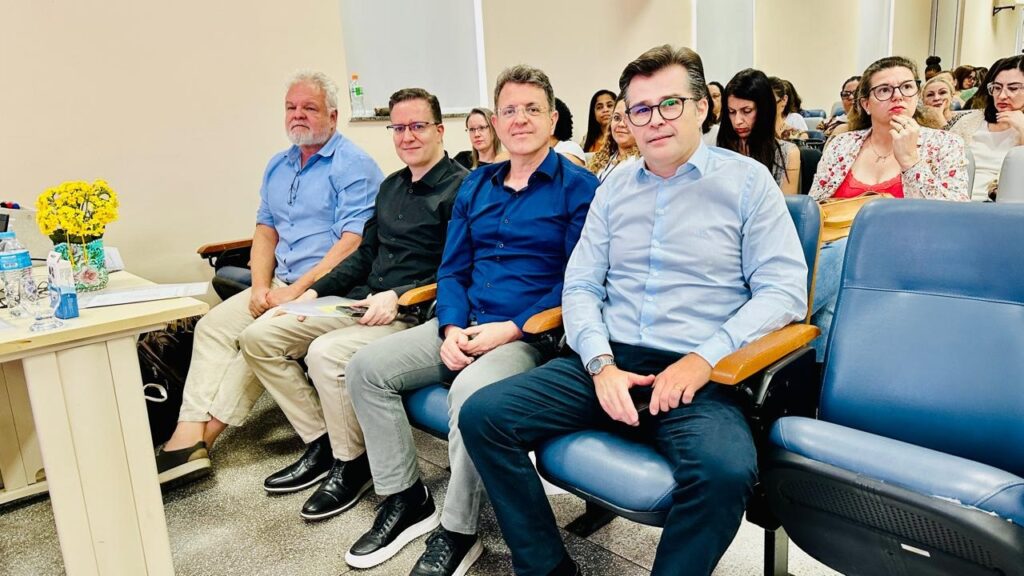 A imagem mostra um grupo de pessoas sentadas em cadeiras em uma sala. Há quatro homens na frente, todos sentados em cadeiras azuis. O homem mais à esquerda tem cabelo grisalho e usa uma camisa azul clara e calças bege. Ao lado dele, um homem com cabelo escuro e óculos veste uma camisa preta e calças cinzas. O próximo homem, também com cabelo escuro e óculos, usa uma camisa azul escura e calças cinzas. O homem mais à direita tem cabelo escuro e usa óculos, vestindo uma camisa azul clara e calças escuras. Atrás deles, um grupo de mulheres está sentado em cadeiras, algumas olhando para a frente e outras parecendo engajadas em conversas. Uma mesa com um vaso de flores amarelas e garrafas de água está posicionada à esquerda da imagem. A sala parece ser um local de reunião ou conferência.