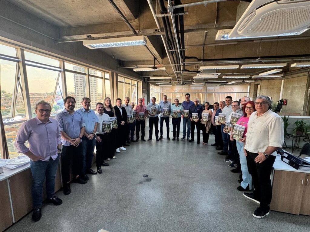 O grupo de pessoas está em pé, alinhado em duas fileiras, em um corredor com grandes janelas ao fundo. A maioria das pessoas está segurando uma publicação semelhante, com a capa voltada para a câmera. A iluminação vem principalmente das janelas, criando um efeito de luz natural. O ambiente parece ser um escritório ou uma sala de reuniões, com mesas e cadeiras visíveis nas laterais e ao fundo. A arquitetura do local é moderna, com tetos altos e luminárias embutidas. As pessoas vestem roupas casuais e formais, indicando uma ocasião profissional. A pose e a forma como seguram as publicações sugerem que estão apresentando ou recebendo algo importante.