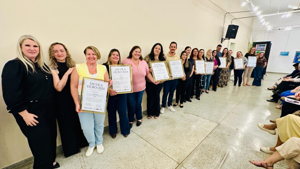 A imagem mostra um grupo de pessoas, principalmente mulheres, em pé em uma fila segurando certificados emoldurados. Os certificados têm o texto "ESCOLA OURO/2024" impresso neles. Elas parecem estar em uma cerimônia de premiação ou evento semelhante, possivelmente em uma escola ou instituição educacional, como sugerido pelo texto do certificado. As pessoas estão vestidas de forma casual e elegante. Ao fundo, há algumas pinturas em exibição e luminárias, indicando um local interno como uma galeria ou salão. Algumas pessoas também estão sentadas ao fundo, observando o evento. A atmosfera geral parece ser de celebração.