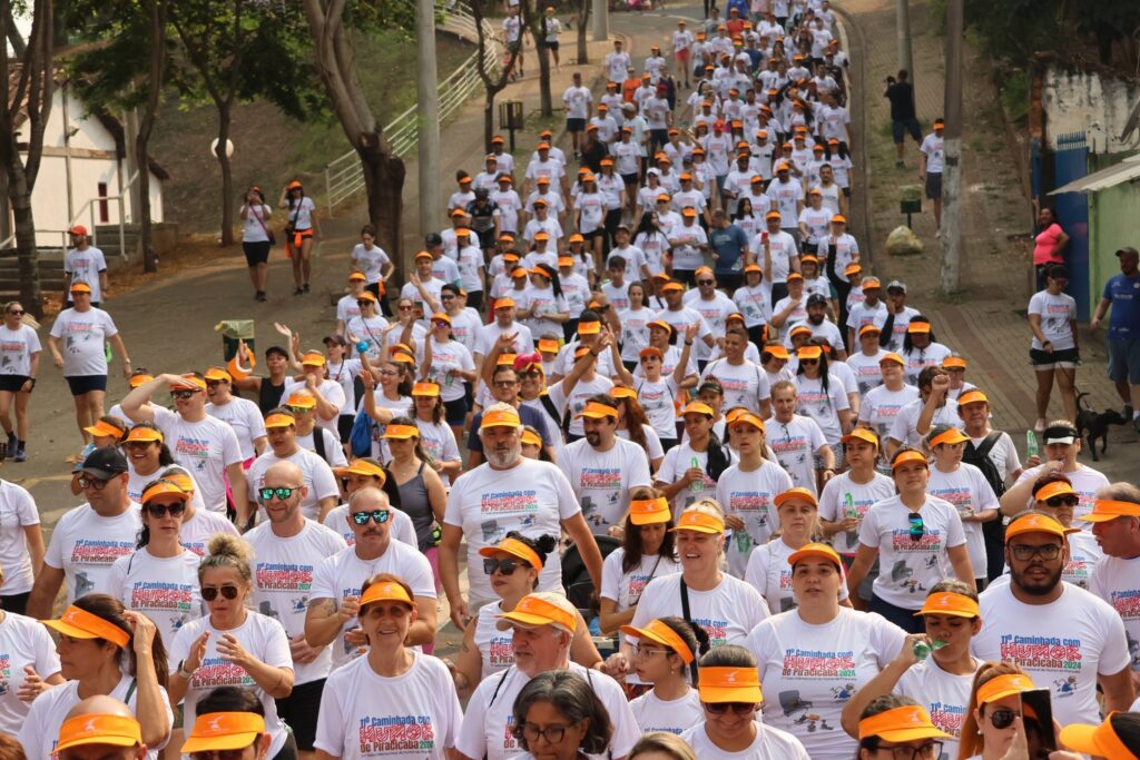 A imagem mostra um grande grupo de pessoas participando de uma caminhada, a maioria vestindo camisetas brancas e bonés ou viseiras laranja. O evento parece ser uma corrida ou caminhada comunitária, possivelmente com fins beneficentes ou de conscientização, como sugerido pelo texto nas camisetas que mencionam "Caminhada com Humor de Piracicaba 2024". Há árvores e uma encosta ao fundo, com algumas pessoas assistindo do lado. O clima parece ser ensolarado, com a maioria das pessoas parecendo animadas e engajadas.
