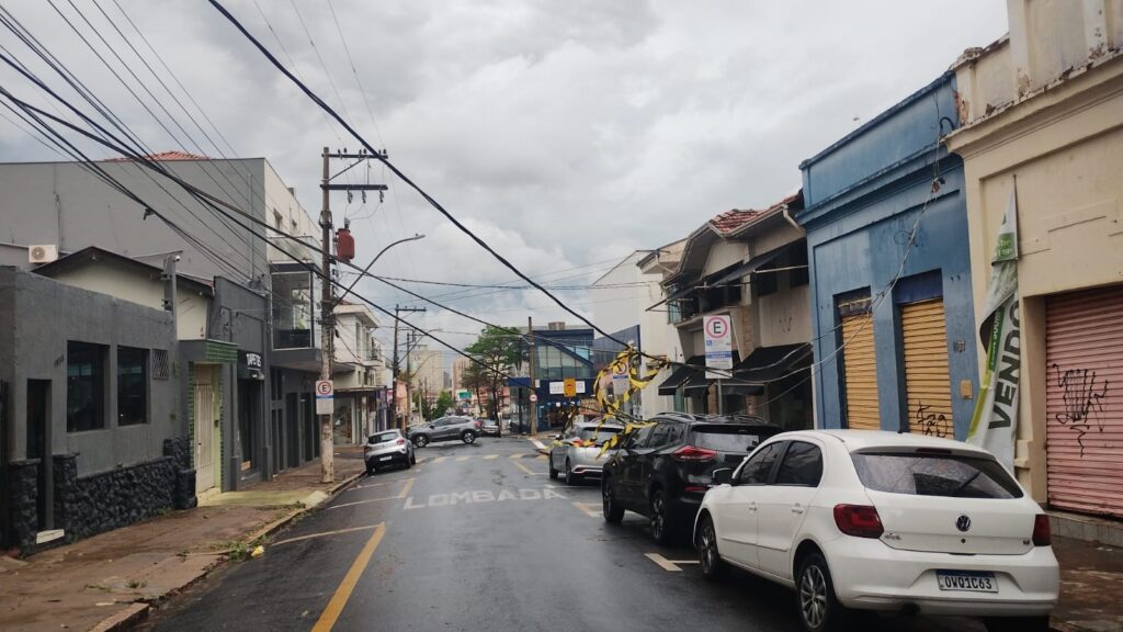 A imagem mostra uma rua com prédios em ambos os lados, com uma linha de carros estacionados. O céu está nublado, e a rua parece estar molhada, sugerindo que choveu recentemente. Há muitos fios elétricos suspensos sobre a rua, que parecem estar em mau estado, com alguns fios caídos ou pendurados perigosamente. Em destaque, um dos fios parece estar preso a um poste e desce em direção à rua, com fitas amarelas e pretas amarradas a ele, possivelmente indicando um perigo ou uma área isolada. Os prédios na rua variam em estilo e cor, com alguns tendo fachadas mais modernas e outros com arquitetura mais antiga. Há sinais de comércio nas fachadas, como lojas e placas. A rua está sinalizada com faixas amarelas no asfalto e uma placa indicando "LOMBADA".