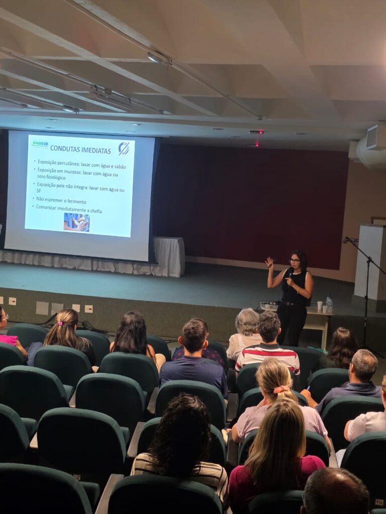 A imagem mostra uma sala de conferências com uma plateia sentada em cadeiras voltadas para um palco. No palco, uma mulher está de pé, falando para a plateia e gesticulando com a mão. Atrás dela, há uma tela de projeção com texto em português. A plateia é composta por pessoas de diversas idades, a maioria delas olhando para a mulher no palco. A iluminação da sala é suave, e a atmosfera parece ser de uma apresentação ou palestra. A tela de projeção exibe o título "CONDUTAS IMEDIATAS" e lista algumas ações a serem tomadas em diferentes tipos de exposição: * Exposição percutânea: lavar com água e sabão * Exposição em mucosas: lavar com água ou soro fisiológico * Exposição pele não integra: lavar com água ou SF * Não espremer o ferimento * Comunicar imediatamente a chefia A presença de um logo "PIRA" na tela sugere que a apresentação é realizada por essa organização.
