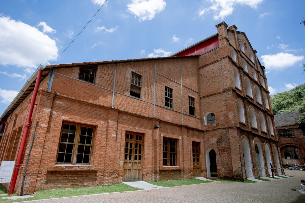 A imagem exibe um edifício histórico de tijolos aparentes, com uma arquitetura industrial marcante. A construção é composta por vários andares e apresenta uma série de janelas de madeira, algumas com vidros quebrados, sugerindo um estado de conservação que mescla preservação e sinais do tempo. A fachada principal é caracterizada por arcos emoldurados em branco, que se repetem em vários níveis, conferindo um ritmo visual à edificação. Em contraste, uma parte do edifício, mais baixa e com um telhado inclinado, possui janelas retangulares e portas de madeira mais simples. Um tubo de drenagem vermelho se destaca na lateral, adicionando um toque de cor à estrutura. O céu azul com nuvens brancas ao fundo e a vegetação verde que emerge ao lado do edifício criam um cenário natural que contrasta com a solidez da construção. O chão é pavimentado com pedras em um padrão de espinha de peixe, e há gramado em algumas áreas próximas às entradas. Há também a presença de algumas pessoas e motocicletas em segundo plano, indicando que o local é acessível e possivelmente um espaço cultural ou de lazer.