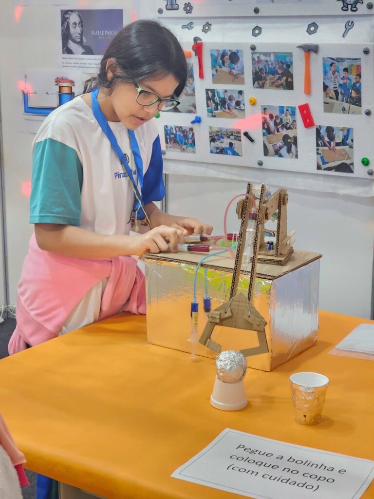 A jovem, usando óculos, está concentrada em um projeto de ciências. Ela usa uma camiseta branca com detalhes em azul e verde e um crachá azul pendurado no pescoço. Ela está manuseando um dispositivo mecânico feito de papelão, que parece ser um braço robótico com pinça, operado por seringas. O dispositivo está montado sobre uma caixa coberta com papel alumínio. No fundo, há um quadro com fotos de crianças trabalhando em projetos e uma imagem de Blaise Pascal. Uma placa no centro da mesa com a inscrição "Pegue a bolinha e coloque no copo (com cuidado)" sugere a tarefa que a jovem está realizando.