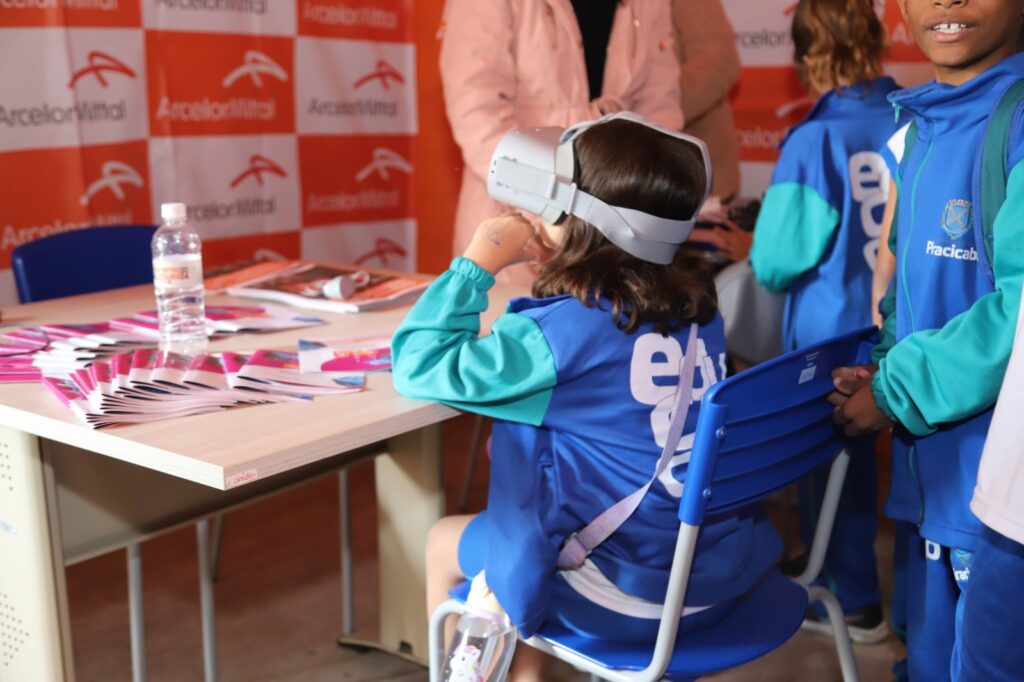 A imagem mostra um grupo de crianças em um evento, possivelmente uma feira de ciências ou uma exposição educacional. Uma menina, sentada em uma cadeira azul, usa um dispositivo de realidade virtual na cabeça. Ela está vestindo uma jaqueta azul com detalhes em verde-água e um logotipo branco com letras roxas em suas costas. Na mesa à sua frente, há uma garrafa de água e várias brochuras ou folhetos empilhados. Ao fundo, um banner com o logotipo "ArcelorMittal" em laranja e branco sugere que a empresa é a patrocinadora ou organizadora do evento. Outras crianças, também vestindo uniformes semelhantes, estão presentes, indicando um ambiente escolar ou de grupo. Uma das crianças, à direita, usa uma jaqueta com a palavra "Praticab" escrita.