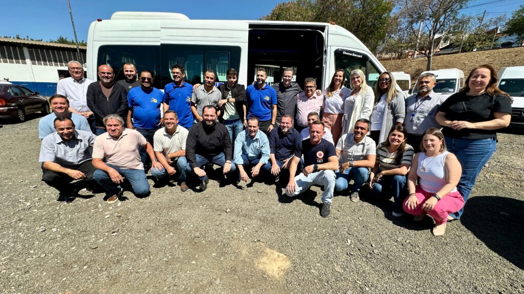 A imagem mostra um grupo de pessoas em frente a uma van branca. A maioria das pessoas está em pé, mas algumas estão agachadas. Todos estão sorrindo e parecem estar em um evento ao ar livre. As pessoas estão vestindo roupas casuais e a maioria são homens. Há algumas mulheres no grupo também. O fundo mostra uma construção, árvores e um céu azul claro. A van parece ser um modelo de transporte de passageiros.