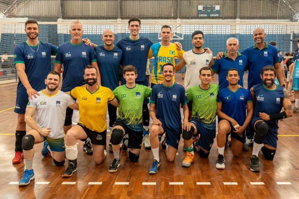 A imagem mostra um grupo de 14 homens, possivelmente uma equipe de vôlei, posando para uma foto em uma quadra. Eles estão vestindo uniformes de time, com vários deles usando camisetas azuis escuras com o logo "BOLSA ATLETA" e um escudo com as iniciais "CBDS". Alguns jogadores usam camisetas amarelas, brancas ou verdes com designs semelhantes. A maioria dos homens está em pé, com alguns ajoelhados na frente. Todos parecem estar em boa forma física e sorrindo para a câmera. O fundo da imagem mostra uma quadra de vôlei com uma rede e arquibancadas ao fundo. A iluminação é clara, sugerindo que a foto foi tirada durante o dia.