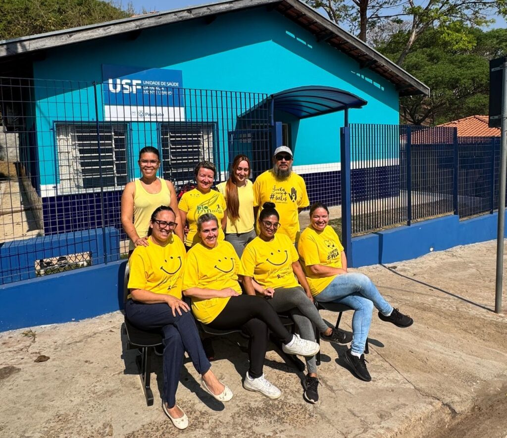 A imagem mostra um grupo de oito pessoas, todos vestindo camisetas amarelas, em frente a um edifício azul claro. O edifício tem uma placa com o texto "USF UNIDADE DE SAÚDE". As pessoas estão em diferentes posições, algumas sentadas em bancos pretos e outras em pé. Todas parecem estar sorrindo e posando para a foto. Há uma grade azul na frente do edifício e algumas árvores e vegetação ao fundo. O dia parece ensolarado.