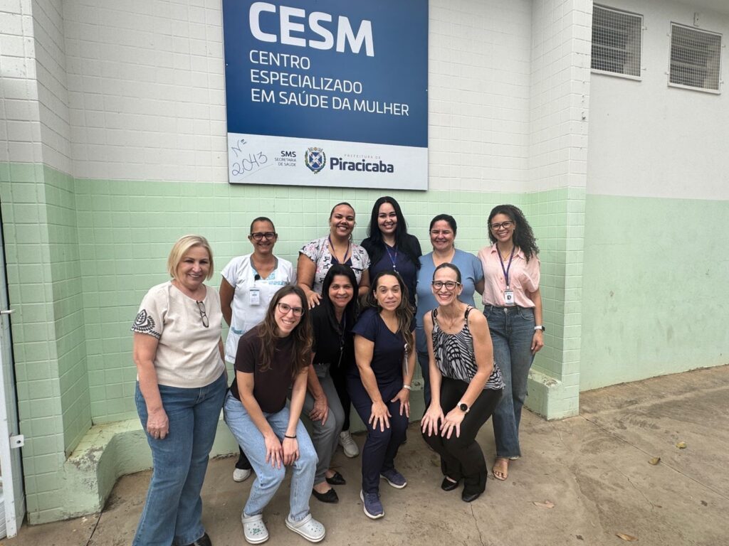 A imagem mostra um grupo de dez mulheres posando em frente a um prédio com uma placa azul e branca que diz "CESM CENTRO ESPECIALIZADO EM SAÚDE DA MULHER". Há também o logotipo da prefeitura de Piracicaba e da Secretaria de Saúde. As mulheres estão vestidas com roupas casuais e profissionais, e a maioria delas está sorrindo para a câmera. Algumas estão em pé e outras estão agachadas ou sentadas. O prédio tem paredes verdes e brancas, e há janelas com grades na parte superior. O chão é de concreto.