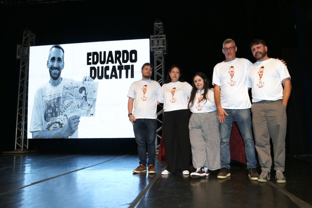 Cinco pessoas, todas usando camisetas brancas estampadas com a caricatura de Eduardo Ducatti, posam no palco em frente a um telão que exibe a foto e o nome do homenageado.