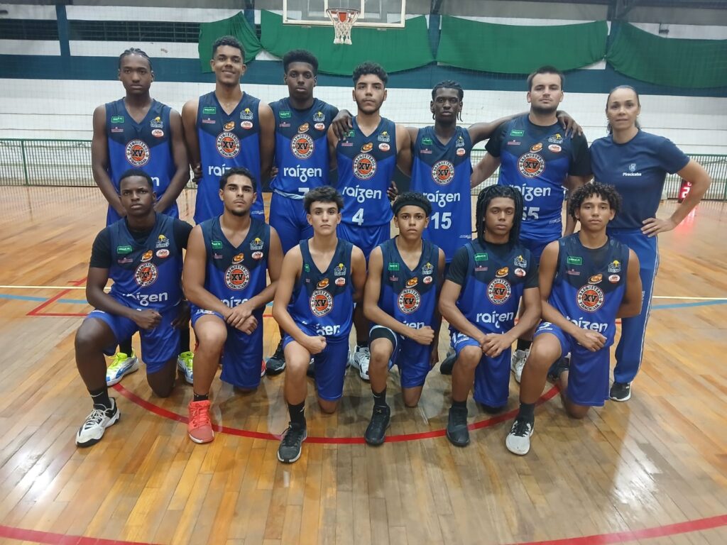 A imagem mostra uma equipe de basquete posando para uma foto em uma quadra de basquete. Há 12 pessoas na foto, incluindo 10 jogadores e 2 treinadores. Todos os jogadores estão vestindo uniformes de basquete azuis com o logotipo "Raizen" e o número "XV" neles. Os jogadores estão em duas fileiras, com cinco jogadores na fileira da frente e cinco na fileira de trás. Os dois treinadores estão atrás dos jogadores. A quadra de basquete tem piso de madeira e uma cesta de basquete ao fundo. As paredes da quadra são verdes e brancas.
