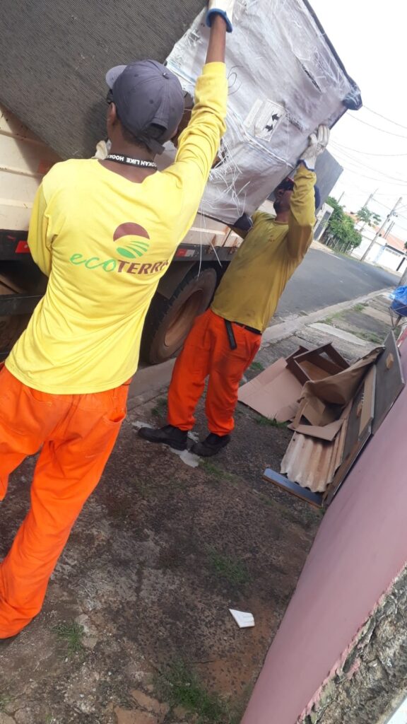 A imagem mostra dois trabalhadores em um dia claro, em frente a um caminhão. Ambos vestem uniformes de trabalho: um com uma camiseta amarela de manga comprida com o logo "ecoterra" nas costas e calças laranja vibrantes; o outro, com uma camiseta amarela de manga comprida e calças laranja. O trabalhador da esquerda, visto de costas, usa um boné escuro e luvas brancas, enquanto segura uma grande peça de material, possivelmente um painel ou cobertura, que está envolta em plástico. O trabalhador da direita, também de luvas brancas, ajuda a carregar o mesmo item. Ambos parecem estar empenhados na tarefa de carregar ou descarregar o material no caminhão. No chão ao redor deles, há vários objetos de descarte, incluindo pedaços de papelão, um pedaço de telha ondulada e outros detritos, sugerindo que estão envolvidos em um trabalho de coleta ou descarte de resíduos. O ambiente parece ser uma área urbana, com uma rua e casas visíveis ao fundo.