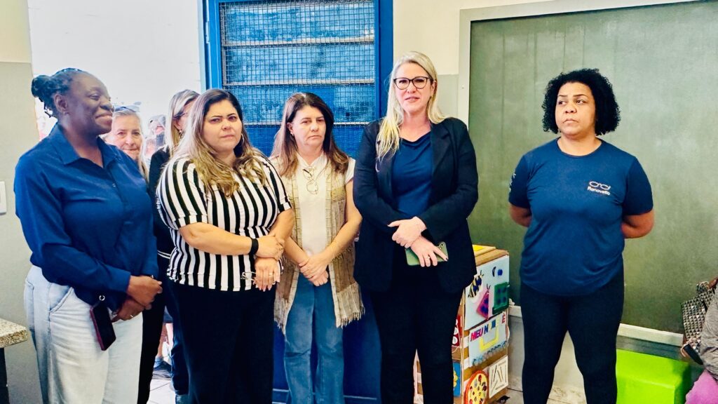 A imagem mostra um grupo de seis mulheres em pé, em um ambiente interno que parece ser uma sala de aula ou um escritório. Da esquerda para a direita: 1. A primeira mulher, à esquerda, tem pele escura, cabelo preto preso em um coque e veste uma camisa azul escura de botões e calças claras. Ela está olhando para a esquerda, com um leve sorriso. Um celular está preso à sua calça. 2. A segunda mulher, atrás da primeira, tem pele clara e cabelo loiro. Ela está sorrindo e olhando para frente. 3. A terceira mulher tem pele clara, cabelo castanho ondulado e veste uma blusa listrada em preto e branco e uma saia preta. Ela está olhando para a frente com uma expressão neutra. Ela segura um par de óculos na mão direita. 4. A quarta mulher tem pele clara, cabelo castanho escuro e veste uma blusa branca e um colete bege com franjas. Ela está olhando para a frente com uma expressão séria.