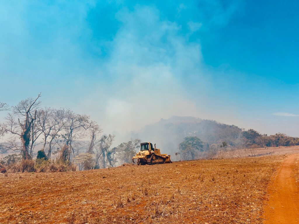 A imagem mostra uma paisagem desolada com um céu azul claro e algumas nuvens de fumaça. No centro da imagem, um trator amarelo está posicionado em uma colina de terra avermelhada e seca, com alguns arbustos e árvores esparsas ao redor. A vegetação parece ter sido queimada ou está em processo de queima, evidenciada pela densa fumaça que se espalha pelo fundo, obscurecendo as colinas distantes. À direita, uma estrada de terra batida sobe a colina. A cena geral transmite uma sensação de destruição ambiental e atividade de desmatamento ou controle de fogo.