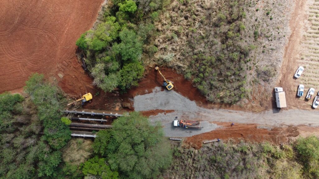 A imagem, tirada de cima, mostra um canteiro de obras em uma área rural. Uma escavadeira amarela está trabalhando em uma área de terra vermelha, perto de tubulações grandes que parecem ser de água ou esgoto. Há também um caminhão com uma plataforma de carga e vários carros estacionados em uma estrada de terra batida. A vegetação ao redor é uma mistura de árvores verdes e arbustos secos, e ao fundo, à direita, é possível ver um campo de plantação. O cenário geral sugere um trabalho de infraestrutura em andamento em um ambiente natural.