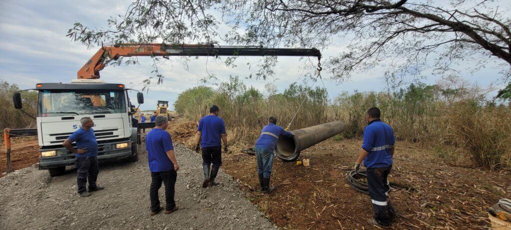 Um caminhão com um guindaste laranja está posicionado em uma estrada de cascalho. Há cinco homens no local, todos vestindo roupas de trabalho azuis. Um dos homens está em pé perto da parte traseira do caminhão, com as mãos nos quadris, olhando para a direita. Outro homem está em pé perto da frente do caminhão, olhando para a esquerda. Há um grande tubo de concreto no chão à direita do caminhão, e dois homens estão perto dele. Há uma pá carregadeira amarela em segundo plano. O céu está nublado.