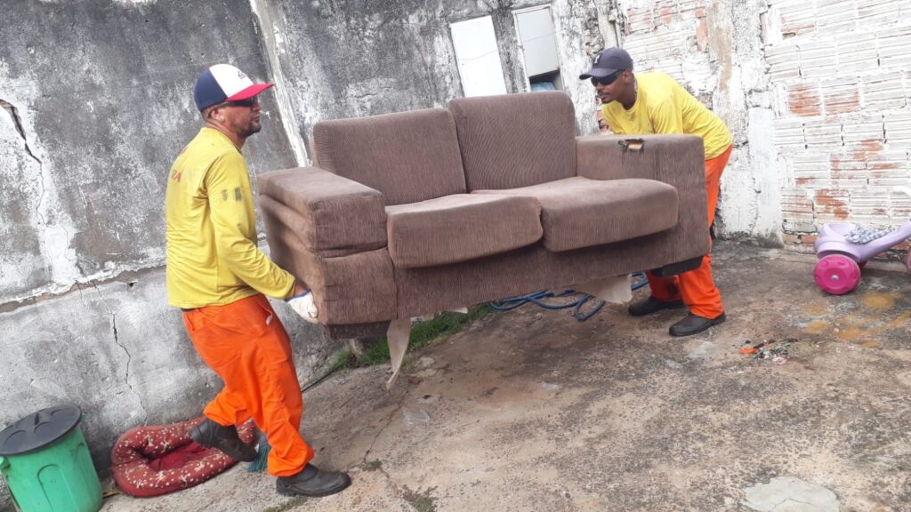A imagem mostra dois homens carregando um sofá marrom em um ambiente externo, possivelmente um quintal. Ambos vestem uniformes de trabalho, com um deles usando uma camiseta amarela e calças laranjas, e o outro também com camiseta amarela e calças laranjas, além de um boné azul e branco. O sofá parece ser de dois lugares e está sendo levantado do chão. No chão, próximo ao sofá, há uma cama de cachorro vermelha com estampa de bolinhas brancas e um balde verde com tampa preta. Ao fundo, observa-se uma parede de concreto texturizada e uma parede de tijolos. Há também um brinquedo infantil com rodas coloridas, um triciclo rosa e roxo, parcialmente visível no lado direito da imagem. O chão é de concreto irregular, com algumas manchas e detritos espalhados. A iluminação sugere que o dia está claro.