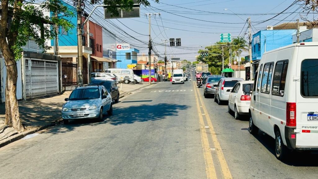 A imagem mostra uma rua movimentada em um dia ensolarado. Vários carros e uma van branca estão estacionados ou em movimento na via. Edifícios coloridos, como um azul e um laranja, ladeiam a rua, com algumas lojas e fachadas visíveis. Fios elétricos e postes de luz cruzam o céu, indicando uma área urbana. Uma árvore com folhagem verde proeminente está à esquerda, projetando sombra na calçada. O tráfego parece moderado, com alguns veículos em movimento e outros parados. Há sinalização de trânsito, incluindo um sinal de "RETORNO" verde e um semáforo ao longe. A estrada é pavimentada e marcada com linhas amarelas duplas no centro.