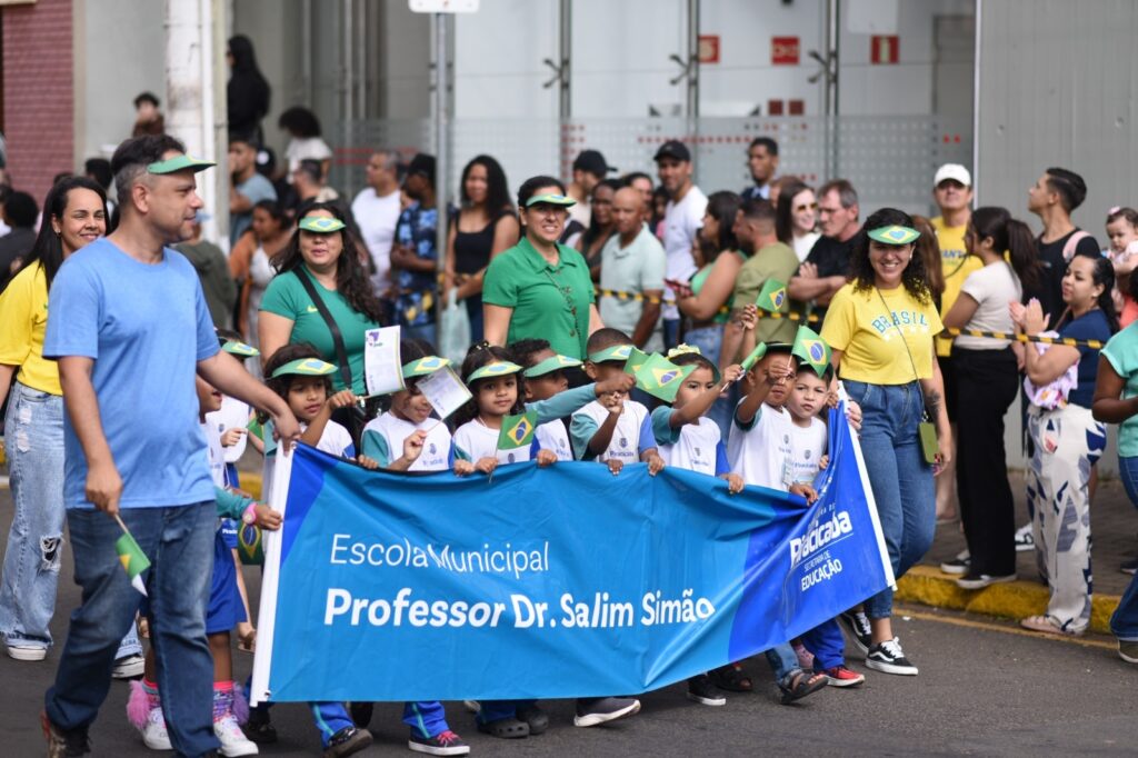 A imagem mostra um desfile escolar, onde crianças vestidas com uniformes brancos e azuis seguram bandeiras do Brasil e um grande banner azul com o texto "Escola Municipal Professor Dr. Salim Simão". Vários adultos acompanham as crianças, alguns com bonés e camisetas com as cores do Brasil, demonstrando apoio e orgulho. O evento parece ser uma celebração cívica ou um evento escolar importante.