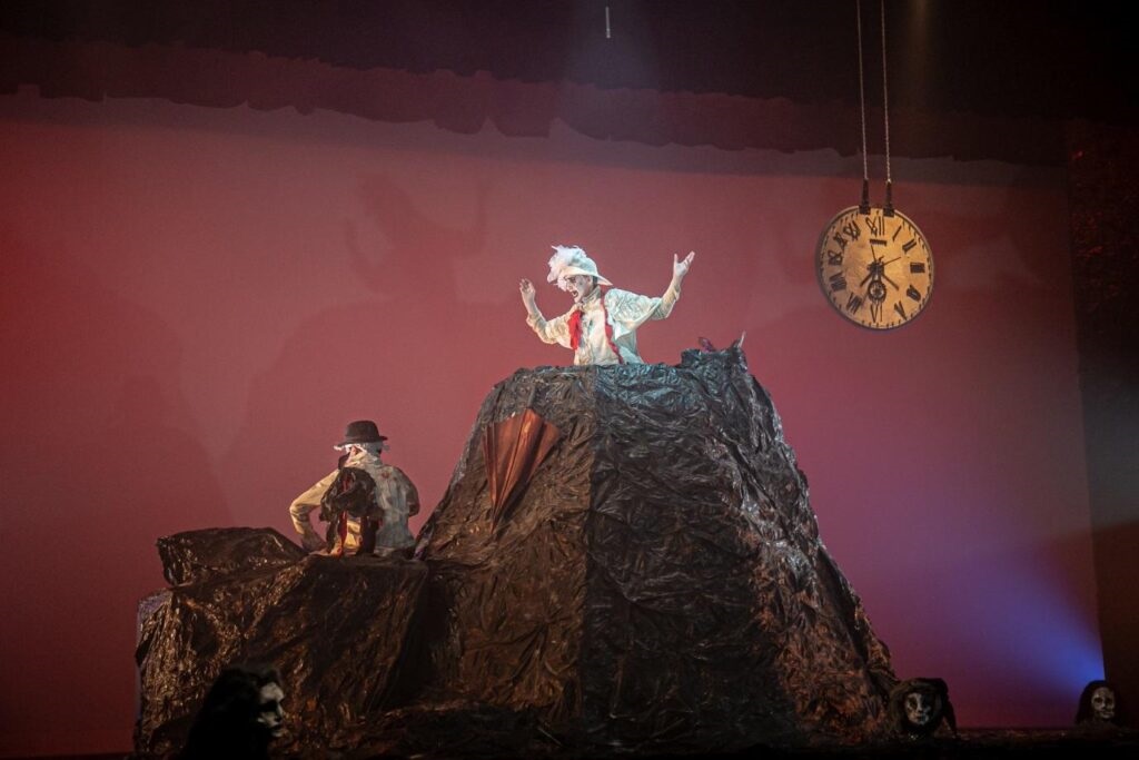 A imagem mostra uma cena teatral com dois personagens vestidos com figurinos elaborados e maquiagem de palhaço, em cima de uma estrutura que se assemelha a uma montanha escura e irregular. O personagem principal, no topo da estrutura, veste roupas brancas com detalhes vermelhos e um chapéu emplumado, com os braços levantados em um gesto dramático. Ele parece estar no centro das atenções. À esquerda, um segundo personagem, também com maquiagem de palhaço e vestindo um chapéu preto, está sentado na estrutura, de costas para o espectador, olhando para o lado. Ao fundo, um grande relógio de parede com números romanos está pendurado, sugerindo uma ênfase no tempo ou em um evento iminente. O cenário é completado por um fundo vermelho vibrante e iluminação teatral que cria sombras e realça os elementos da cena. Há também o que parecem ser rostos ou crânios decorando a base da estrutura, adicionando um tom sombrio e teatral à apresentação.