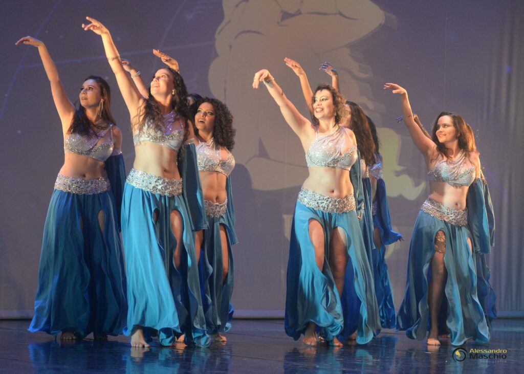 A imagem mostra um grupo de dançarinas do ventre em um palco, vestindo trajes elaborados. Elas usam tops curtos adornados com pedras brilhantes e saias longas e fluidas em tons de azul claro, com fendas altas que revelam as pernas. Os cintos brilhantes na cintura complementam os trajes. As dançarinas estão em poses graciosas, com os braços levantados e expressões focadas. O fundo é escuro, com projeções abstratas em tons claros, e o palco apresenta um reflexo brilhante no chão. A iluminação foca nas dançarinas, destacando seus movimentos e os detalhes de suas vestimentas. Uma assinatura de fotógrafo, "Alessandro Maschio fotografia", aparece no canto inferior direito.